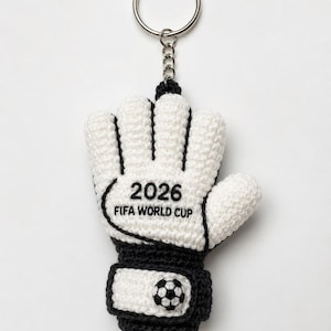 以下が含まれることがあります： 白と黒のサッカーゴールキーパーグローブの形をしたかぎ針編みのキーホルダー。グローブには「2026 FIFA WORLD CUP」の文字とサッカーボールのグラフィックが入っています。キーホルダーにはシルバーのリングが付いています。