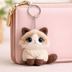 May include: A crochet cat keyring with a silver ring. The cat has brown ears, paws, and tail, with a cream-coloured body. It features large blue eyes and pink cheeks. The keyring is shown next to a pink purse.