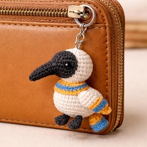 Peut inclure: Porte-clés en forme d'oiseau crocheté, avec un bec, une tête et des pieds noirs, et un corps blanc. L'oiseau a un col et des accents d'ailes rayés jaune et bleu. Le porte-clés est attaché à un portefeuille en cuir marron.