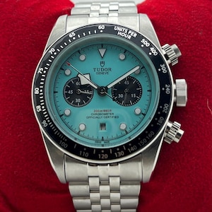 Op de afbeelding: Een Tudor chronograaf horloge van roestvrij staal met een heldere turquoise wijzerplaat, zwarte sub-wijzerplaten en een zwarte tachymeter ring. Het horloge heeft een datumvenster op de zes-uur positie, lichtgevende indexen en een schakelarmband.