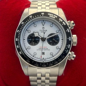 May include: A silver-tone stainless steel chronograph watch with a white dial, two black sub-dials, and a black tachymeter bezel. The watch features a date window at the six o'clock position, luminous hour markers, and a metal link bracelet strap.