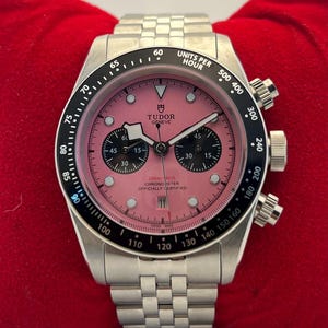 Peut inclure: Une montre chronographe en acier inoxydable avec un cadran rose, des sous-cadrans noirs et une lunette tachymètre noire. La montre présente un bracelet à maillons argentés, des index lumineux et le texte Tudor Geneve, 200m:660ft, Chronometer Officially Certified, Swiss Made.