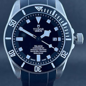 Peut inclure: Une montre-bracelet noire et argentée avec un cadran noir et un bracelet en caoutchouc noir. Le cadran de la montre comporte des marqueurs blancs, des aiguilles et le texte "TUDOR GENEVE PELAGOS CHRONOMETER". La montre est étanche à 500m.