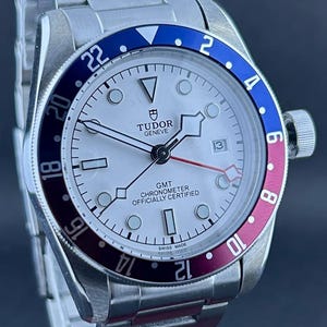 May include: A stainless steel Tudor Geneve GMT watch with a white face and a blue and burgundy rotating bezel. The watch has a date display at 3 o'clock and a stainless steel strap.