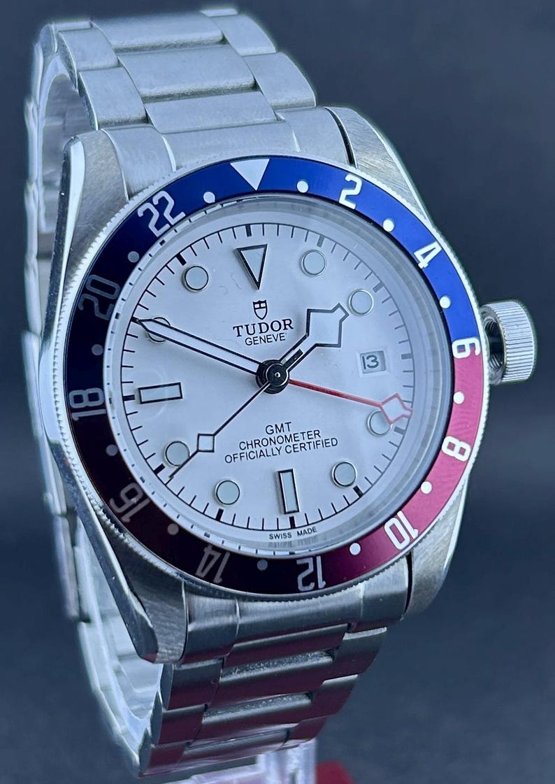 May include: A stainless steel Tudor Geneve GMT watch with a white face and a blue and burgundy rotating bezel. The watch has a date display at 3 o'clock and a stainless steel strap.