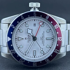 May include: A stainless steel wristwatch with a white face, featuring a red and blue bezel. The watch has a silver strap and a date display at the 3 o'clock position. The face includes the text "TUDOR GENEVE GMT CHRONOMETER OFFICIALLY CERTIFIED".