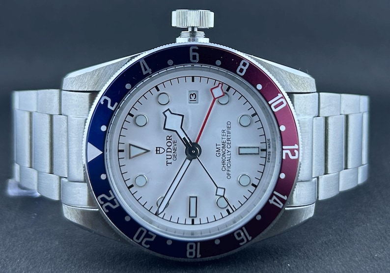 May include: A stainless steel wristwatch with a white face, featuring a red and blue bezel. The watch has a silver strap and a date display at the 3 o'clock position. The face includes the text "TUDOR GENEVE GMT CHRONOMETER OFFICIALLY CERTIFIED".