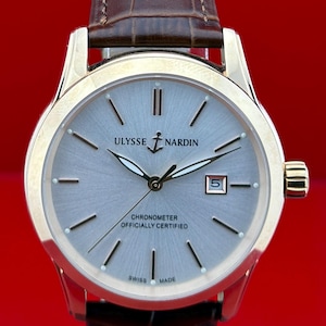 May include: A gold-toned Ulysse Nardin wristwatch with a light gray face and brown leather band. The watch face displays the brand name, the word "Chronometer", and the date. The watch is against a red background.