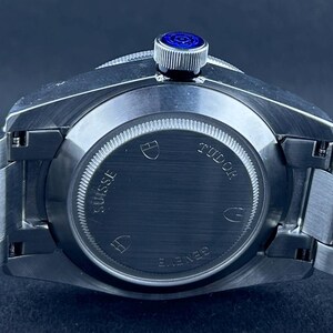 May include: A close-up shot of a stainless steel wristwatch. The watch has a silver strap and a dark blue dial. The back of the watch is visible, with the brand name "TUDOR" and the words "SUISSE" and "GENEVE" displayed.