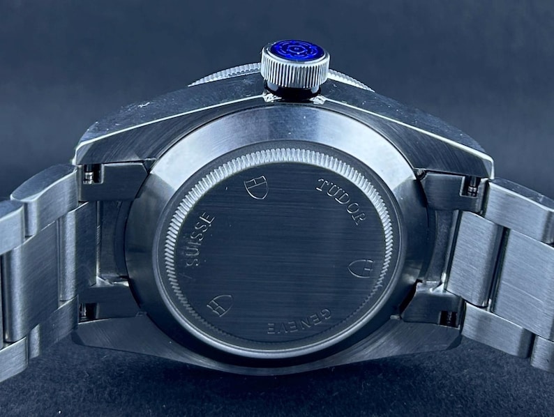 May include: A close-up shot of a stainless steel wristwatch. The watch has a silver strap and a dark blue dial. The back of the watch is visible, with the brand name "TUDOR" and the words "SUISSE" and "GENEVE" displayed.