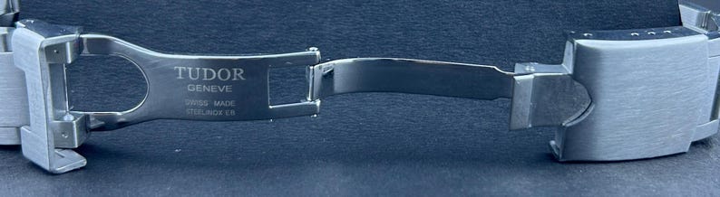 May include: A close-up of a stainless steel watch clasp, with the brand name "TUDOR GENEVE" and "SWISS MADE" text. The clasp is open, showing the inner workings and the brushed metal finish. The watch band is also visible.