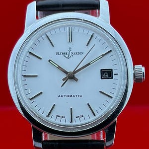May include: A silver Ulysse Nardin automatic wristwatch with a white face, black hands, and a date window. The watch has a black leather strap and a silver crown. The watch face displays the text "Ulysse Nardin" and "Automatic".
