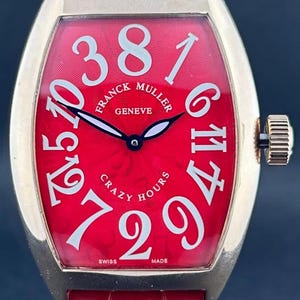 May include: A luxury wristwatch with a red face and a gold-colored case. The watch face features large white numerals and the words "FRANCK MULLER GENEVE CRAZY HOURS". The watch has a red leather band.