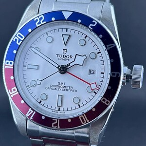 May include: A silver wristwatch with a white face and a blue and red bezel. The watch has a date display at the 3 o'clock position and a stainless steel strap. The watch face displays the text "TUDOR GENEVE" and "GMT CHRONOMETER OFFICIALLY CERTIFIED".