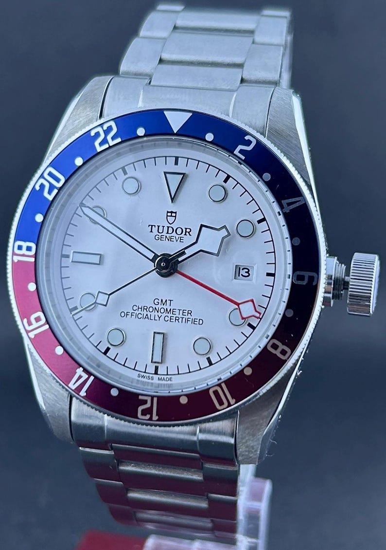 May include: A silver wristwatch with a white face and a blue and red bezel. The watch has a date display at the 3 o'clock position and a stainless steel strap. The watch face displays the text "TUDOR GENEVE" and "GMT CHRONOMETER OFFICIALLY CERTIFIED".