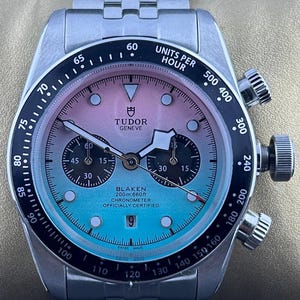 Puede incluir: Reloj de pulsera de acero inoxidable con esfera degradada de rosa a azul. El reloj tiene un bisel negro con marcas blancas, tres subesferas y una ventana de fecha. La correa del reloj también es de acero inoxidable.