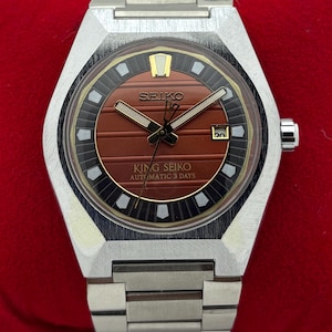 Op de afbeelding: Een zilverkleurig King Seiko automatisch polshorloge met een getextureerde, horizontaal gestreepte roodbruine wijzerplaat, een zwarte buitenring met witte indexen, een datumvenster op de drie-uurpositie en een roestvrijstalen schakelarmband.