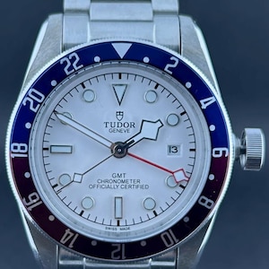 May include: A stainless steel Tudor GMT watch with a white face, a blue and burgundy bezel, and a date display at the 3 o'clock position. The watch has a silver strap and is labelled "GMT Chronometer Officially Certified."