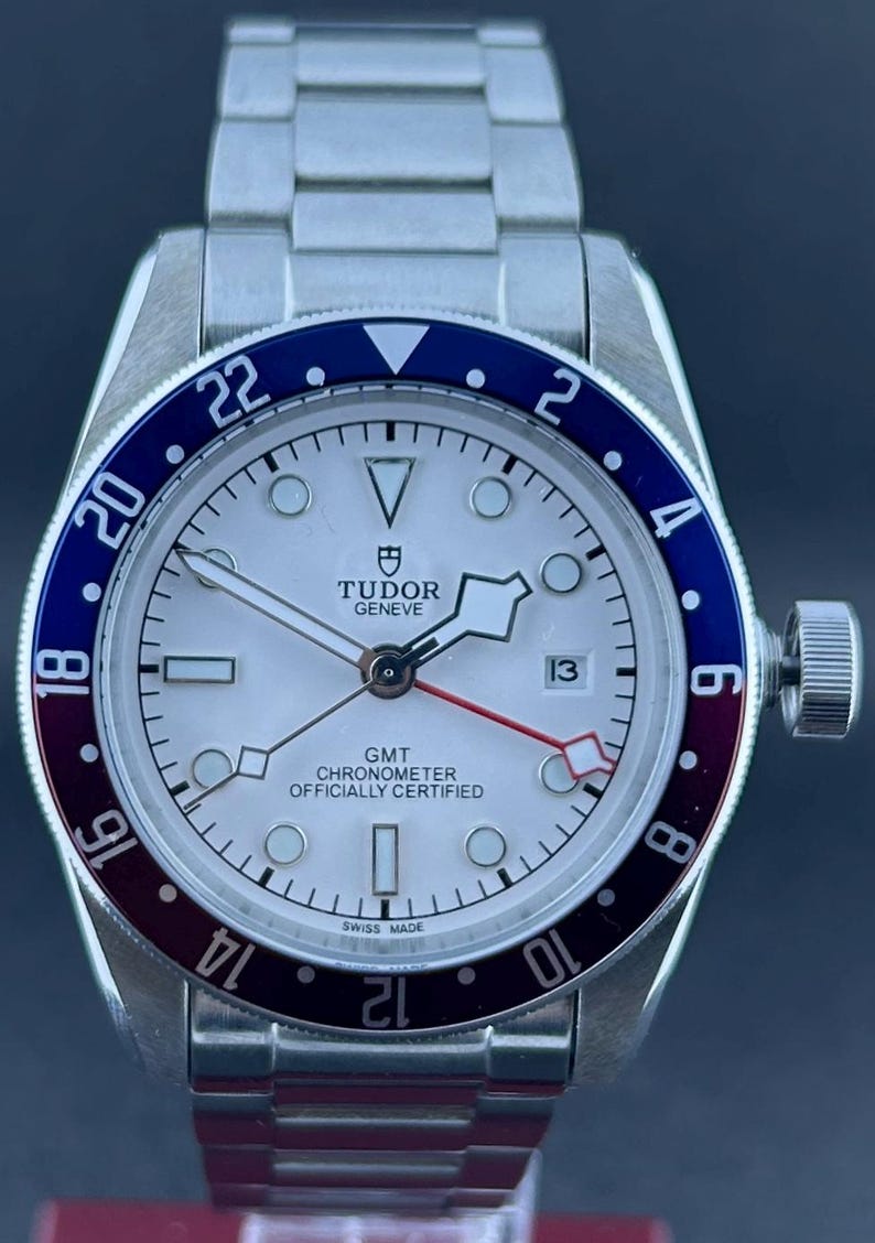 May include: A stainless steel Tudor GMT watch with a white face, a blue and burgundy bezel, and a date display at the 3 o'clock position. The watch has a silver strap and is labelled "GMT Chronometer Officially Certified."