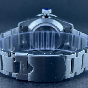 May include: A stainless steel wristwatch with a silver strap and a black clasp. The watch face is not visible, but the crown has a blue accent. The clasp is engraved with the word "TUDOR".