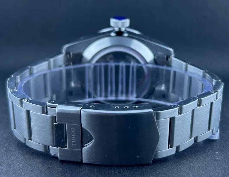May include: A stainless steel wristwatch with a silver strap and a black clasp. The watch face is not visible, but the crown has a blue accent. The clasp is engraved with the word "TUDOR".