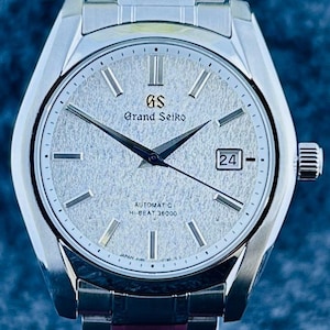 May include: Silver-toned Grand Seiko automatic wristwatch with a textured silver dial, black hands, and a date window at the 3 o'clock position. The watch features the text "Grand Seiko" and "Automatic Hi-Beat 36000". The watch has a silver band.
