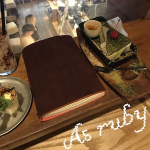 Può includere: Un taccuino A5 in pelle marrone con interno rosso, appoggiato su una superficie di legno. Accanto, un dolce con una ciliegia e una bevanda al cioccolato. Il testo "A5 ruby" è nell'angolo in basso a destra. Il taccuino misura circa 21 cm di altezza.