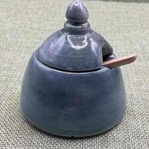 May include: A small, round, grey ceramic jar with a glossy finish and a matching lid featuring a pointed finial. A small, light-coloured wooden spoon is inserted into a notch on the side of the jar, resting against the lid and the rim of the container.