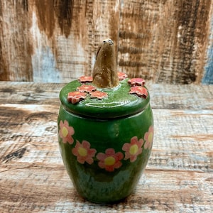 Può includere: Un barattolo in ceramica verde con un coperchio caratterizzato da una figura di coniglio marrone scolpita. Il barattolo e il coperchio sono decorati con fiori rosa dipinti a mano con centri gialli. La superficie della ceramica ha una finitura lucida e il coperchio è rimovibile.