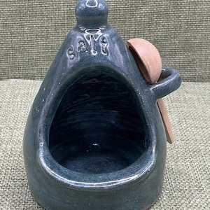 Könnte beinhalten: Ein dunkelblau-graues Salzgefäß aus Keramik mit glänzender Oberfläche und einer breiten Öffnung an der Vorderseite. Das Wort salt ist oben eingeprägt. Ein kleiner Holzlöffel steckt in einem seitlichen Griff und liegt am runden Gefäß an.