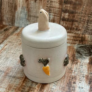 Op de afbeelding: Een lichtroze keramische pot met een deksel, met een kleine konijnenfiguur bovenop. De pot is versierd met een wortel en twee konijnenfiguren. De pot is een decoratief item.