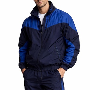 Op de afbeelding: Marineblauwe en koningsblauwe trainingspak. De jas heeft een ritssluiting, een hoge kraag en blauwe panelen op de schouders en mouwen. De bijpassende broek heeft een blauwe streep aan de zijkant. Het model draagt een wit t-shirt.
