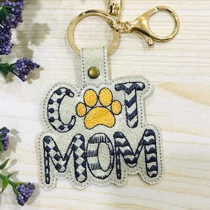 May include: A light gray keychain with the words "CAT MOM" embroidered in black and white patterns. A yellow paw print is in the center. The keychain has a gold-colored ring and clasp.