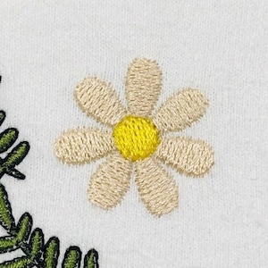 K&ouml;nnte beinhalten: Eine wei&szlig;e Stoffoberfl&auml;che mit gestickten Details. Eine zentrale G&auml;nsebl&uuml;mchenbl&uuml;te mit hellbeigen Bl&uuml;tenbl&auml;ttern und einem leuchtend gelben Zentrum befindet sich neben einem geschwungenen gr&uuml;nen Farnzweig mit detaillierten Stichen.
