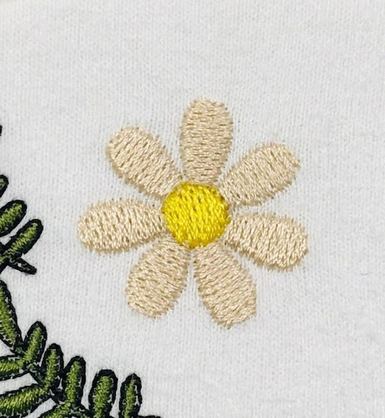 K&ouml;nnte beinhalten: Eine wei&szlig;e Stoffoberfl&auml;che mit gestickten Details. Eine zentrale G&auml;nsebl&uuml;mchenbl&uuml;te mit hellbeigen Bl&uuml;tenbl&auml;ttern und einem leuchtend gelben Zentrum befindet sich neben einem geschwungenen gr&uuml;nen Farnzweig mit detaillierten Stichen.