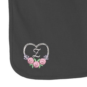 May include: A dark gray baby bib featuring an embroidered design of a white heart outline surrounding the letter Z in white thread, accented with three pink roses with green leaves and small purple floral sprigs at the bottom of the heart.