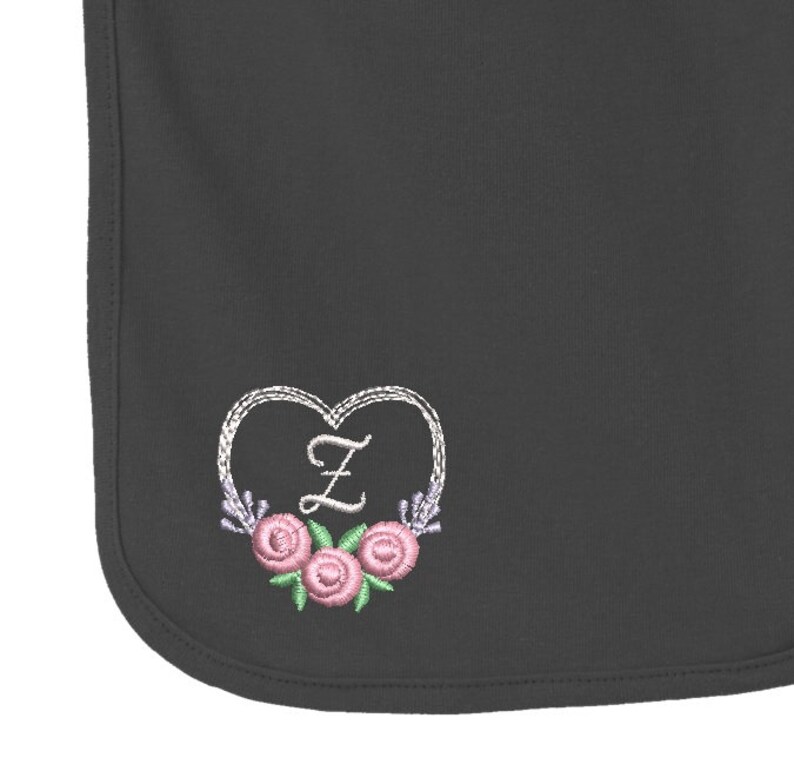 May include: A dark gray baby bib featuring an embroidered design of a white heart outline surrounding the letter Z in white thread, accented with three pink roses with green leaves and small purple floral sprigs at the bottom of the heart.