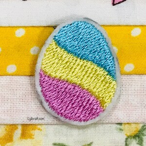 May include: Embroidered egg-shaped patch with blue, yellow, and pink horizontal stripes. The patch is outlined in white and sits on a background of patterned fabric. The text "Lyliacraft.com" is visible.