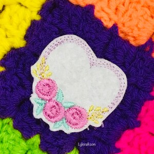 May include: A white felt heart-shaped patch featuring embroidered pink flowers, light green leaves, and yellow accents, with a light purple stitched border. The patch is placed against a vibrant, multi-colored crocheted background with the text Lylicraft.com.