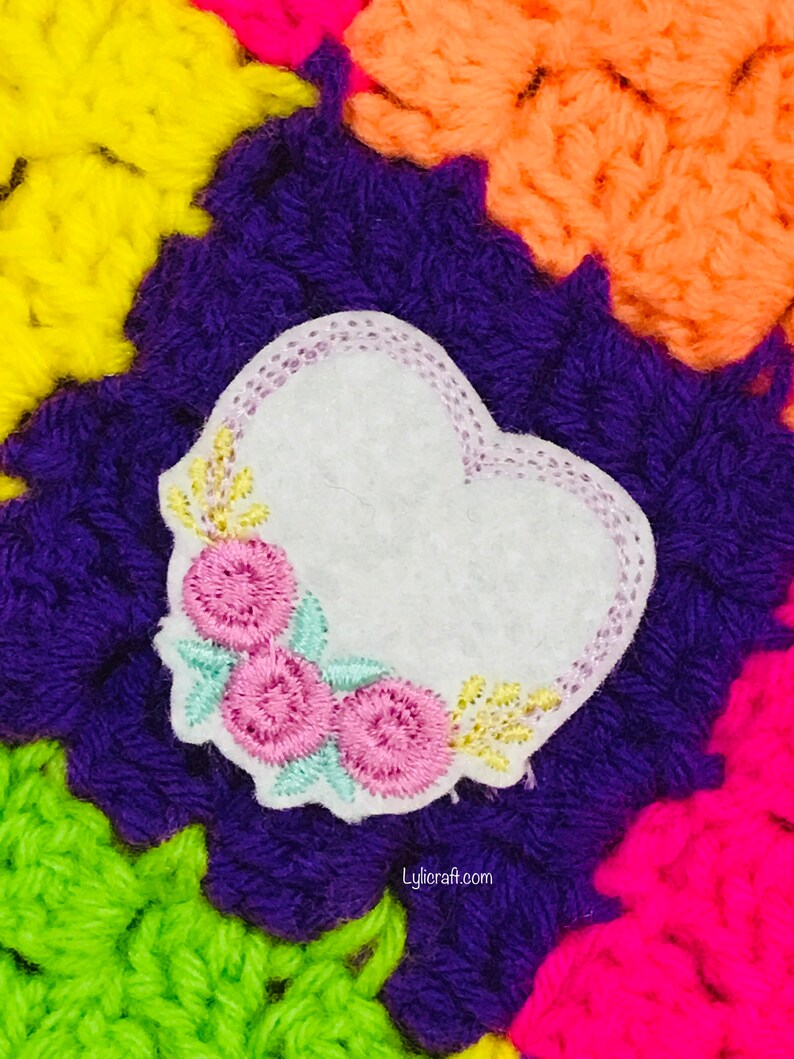 May include: A white felt heart-shaped patch featuring embroidered pink flowers, light green leaves, and yellow accents, with a light purple stitched border. The patch is placed against a vibrant, multi-colored crocheted background with the text Lylicraft.com.