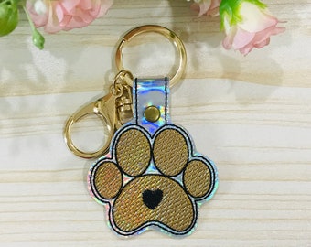 May include: A gold-colored keychain featuring a paw print design with a black heart in the center. The paw print is outlined in black and has a textured appearance. The keychain includes a gold clasp and a gold key ring.