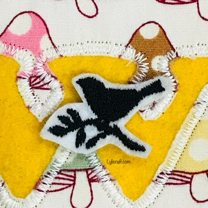 May include: A yellow felt applique with a white stitched border, shaped like a letter W. The applique features a black bird with a white outline and a small branch with leaves, all on a white background with a repeating pattern of pink and brown mushrooms.