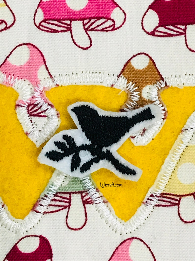 May include: A yellow felt applique with a white stitched border, shaped like a letter W. The applique features a black bird with a white outline and a small branch with leaves, all on a white background with a repeating pattern of pink and brown mushrooms.
