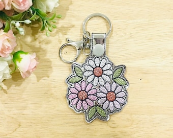 May include: A silver key chain with a charm featuring three embroidered flowers in shades of pink, white, and green.