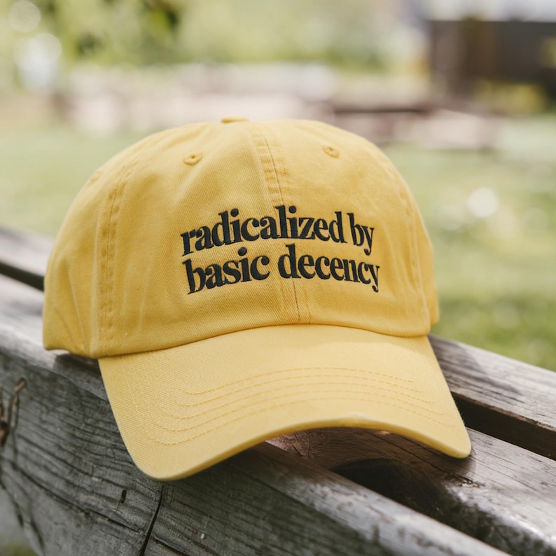 Peut inclure: Casquette de baseball jaune moutarde avec l'inscription "radicalized by basic decency" en noir. La casquette a une visi&egrave;re incurv&eacute;e et est faite d'un tissu doux. Le chapeau est pos&eacute; sur une surface en bois.