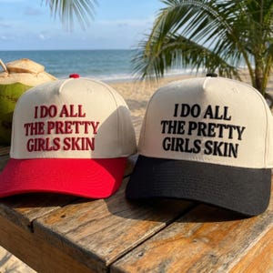 May include: Two baseball caps with the text "I DO ALL THE PRETTY GIRLS SKIN". One cap has a red brim and red text, the other has a black brim and black text. Both caps have a cream-colored crown.