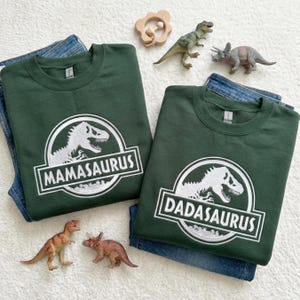 May include: Two dark green sweatshirts with white dinosaur graphics and the words "MAMASAURUS" and "DADASAURUS". The sweatshirts are folded on top of blue jeans. Toy dinosaurs and a wooden toy are also in the image.