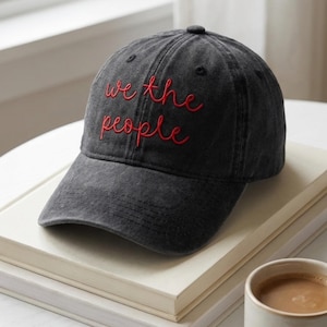 Könnte beinhalten: Dunkelgraue Baseballkappe mit dem gestickten Text "we the people" in Rot. Die Kappe liegt auf einem Stapel Bücher. Eine Tasse Kaffee befindet sich in der unteren rechten Ecke.