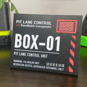 Op de afbeelding: Een zwart rechthoekig bord met witte tekst die PIT LANE CONTROL RaceWatch Transponder, BOX-01 en PIT LANE CONTROL UNIT leest. Het bevat een rood omrande ventilatiegrafiek, rode diagonale waarschuwingsstrepen en de tekst WARNING: FIA SEALED UNIT.