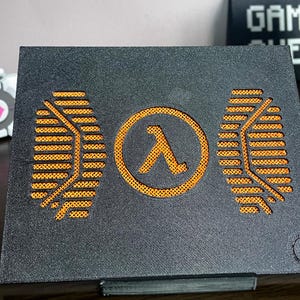 Puede incluir: Un soporte rectangular negro que presenta un recorte circular naranja central con un símbolo lambda griego. Dos patrones texturizados naranjas simétricos flanquean el centro, y un pequeño logotipo circular está grabado en la esquina inferior derecha.